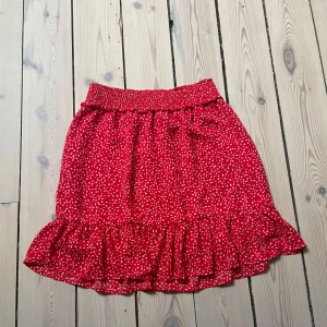 Röd kjol från Holy and Whyte by Lindex  - Röd kjol från Holy and Whyte med vita små hjärtan. Kjolen har en volangdetalj vid fållen och en smockad midja. Perfekt för en somrig look!