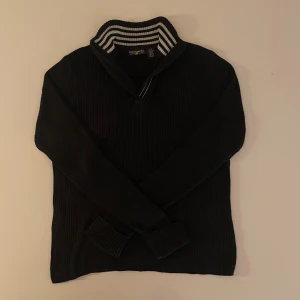 Svart ribbad tröja från Polo Jeans Co. - Säljer en svart ribbad tröja från Polo Jeans Co. med en stilren dragkedja och randig krage i svart och vitt. Tröjan har långa ärmar och en liten RL-logga med amerikansk flagga på kragen. Perfekt för en casual look.