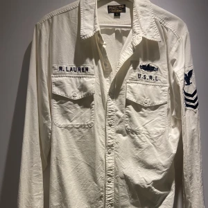 Vit skjorta från Ralph Lauren - Säljer en vit långärmad skjorta från Ralph Lauren med broderade detaljer på bröstet och ärmen. Skjortan har två bröstfickor med knappar och en klassisk krage. Perfekt för en stilren look.