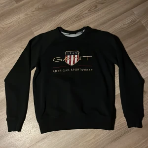 Svart sweatshirt från GANT,    Storlek 13/14 år.   158/164 cm - Säljer en stilren svart sweatshirt från GANT med deras klassiska logga och texten 'American Sportswear' på framsidan. Tröjan har långa ärmar och en rund halsringning. Perfekt för en avslappnad och sportig look.