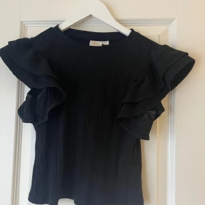 Svart ribbad topp från Gina Tricot - Säljer en svart ribbad topp från Gina Tricot med volangdetaljer på ärmarna. Toppen har en tight passform och korta ärmar.💗