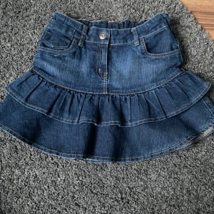 Blå jeanskjol med volang - Säljer en söt blå jeanskjol med volangdetaljer. Kjolen har en klassisk femficksdesign och en knapp framtill. Perfekt för en avslappnad stil. Den är low Waist och skulle säga är Xs, säljer pga används inte💗