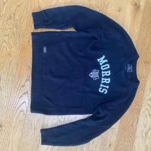Marinblå tröja från Morris - Snygg marinblå tröja från Morris med broderad logga och emblem på bröstet. Tröjan har långa ärmar och ribbade muddar vid ärmslut och nederkant. Perfekt för en stilren look. Inga tecken på användning förutom lite sliten färg men inget man tänker på. Hör av er vid frågor!