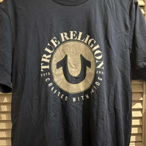 Mörkblå t-shirt från True Religion - Säljer en mörkblå t-shirt från True Religion med ett stort tryck på framsidan. T-shirten har korta ärmar och är tillverkad i bomull. Perfekt för en avslappnad stil.