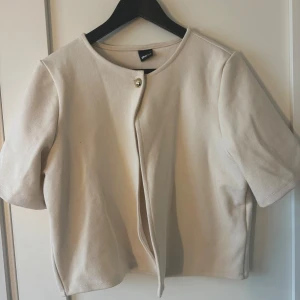 Beige bolerotopp med knapp - Säljer en stilren beige bolerotopp från Tofu med en enkel knapp framtill. Toppen har korta ärmar och en rund halsringning. Perfekt för en elegant look.