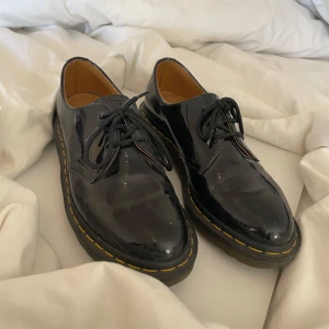Svarta lackskor från Dr. Martens - Snygga svarta lackskor från Dr. Martens med klassisk gul söm. Skorna har snörning och en robust sula, perfekt för en stilren look. Innersulan är märkt med Dr. Martens logotyp.