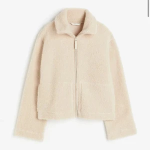 Beige teddyjacka med dragkedja - Mysig beige teddyjacka med dragkedja framtill och två stora fickor. Perfekt för kyliga dagar med sin fluffiga och varma design. Jackan har en hög krage för extra värme och stil.