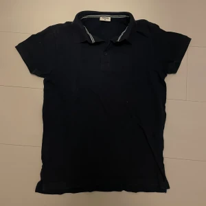 Mörkblå pikétröja från Kappahl - Säljer en klassisk mörkblå pikétröja från Kappahl. Tröjan har korta ärmar och en krage med knappar framtill. Perfekt för en avslappnad stil.