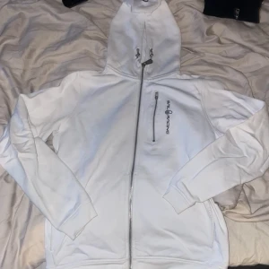 Vit huvtröja från Sail Racing - Säljer en stilren vit huvtröja från Sail Racing med dragkedja och justerbar huva. Tröjan har en bröstficka med dragkedja och logotyp på bröstet. Perfekt för en avslappnad stil. Bröst fickans dragkedja har gått sönder men går fortfarande att öppnas. I storlek 160 som passar storlek S 