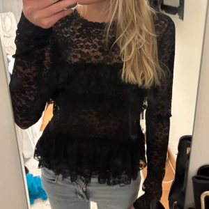 Svart spetsblus med volanger - Snygg svart spetsblus med volanger och långärmad design. Blusen har en elegant knytning i ryggen och är perfekt för en stilfull look. Passar bra till både jeans och kjol. Aldrig använd