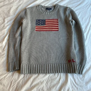 Grå stickad tröja från Ralph Lauren - Säljer en stilren grå stickad tröja från Polo Ralph Lauren med en broderad amerikansk flagga på framsidan. Skick 9.5/10. Perfekt för en avslappnad och klassisk look.