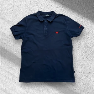 Mörkblå Armani Piké - Snygg mörkblå pikétröja från Emporio Armani med röd broderad logga på bröstet och ärmen. Tröjan har en klassisk krage och knappar framtill. Perfekt för en stilren look. Den är i fint skick. Den passar perfekt att ha under en tjockare tröja eller varför inte bara ha den som den är! Mitt pris är bara 199kr och för följare blir den 189kr vilket är ett superbra pris då nypris på denna ligger runt 1300kr. Jag rekommenderar den starkt för någon som vill ha lite extra stil. Ställ gärna frågor!
