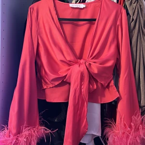 Röd omlottblus med fjäderdetaljer - Trendig röd omlottblus från Glamorous med knytning framtill och fjäderdetaljer vid ärmsluten. 