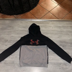 Under Armour - Säljer en snygg svart och grå hoodie från Under Armour med deras logga i rött framtill. Hoodien har en bekväm passform och är perfekt för kyliga dagar. Den har en klassisk design med huva och långa ärmar.