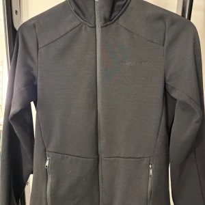 Svart jacka från Peak Performance - Snygg svart jacka från Peak Performance med dragkedja framtill och två praktiska fickor med dragkedjor. Jackan har en stilren design och är perfekt för kyligare dagar. Den är långärmad och har en hög krage för extra värme.