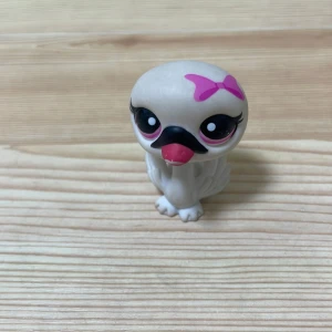 Vit fågel med rosa rosett från Littlest Pet Shop - Söt liten vit fågel från Littlest Pet Shop med stora ögon och en rosa rosett på huvudet. Den har en svart näbb och rosa detaljer runt ögonen. Perfekt för samlare eller som leksak.