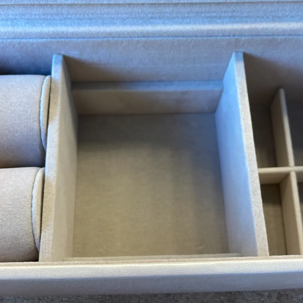 Elegant beige smyckeskrin som aldrig använts med inbyggd spegel i locket. Skrinet har flera fack för förvaring av smycken, inklusive kuddar för armband och ringar. Perfekt för att organisera dina smycken på ett stilfullt sätt. Bredd= 21 cm Höjd= 8,5 cm. Asusteet.