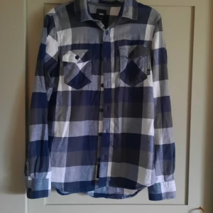 Rutig skjorta från Vans - Snygg rutig flanellskjorta från Vans i blått och vitt. Skjortan har långa ärmar och knappar framtill samt två bröstfickor med knappdetaljer. Står strl XS, skulle jämställa det med strl 164 cirka