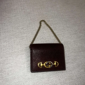Gucci Brun plånbok med guldkedja - Säljer en elegant brun plånbok med en stilren guldkedja och ett snyggt guldfärgat spänne på framsidan. Plånboken har flera fack för kort och en praktisk knappstängning. Perfekt för att hålla ordning på dina kort och kontanter med stil.