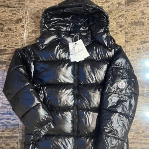 Svart pufferjacka från Moncler(byta) - Snygg svart pufferjacka från Moncler med glansig finish. Jackan har en dragkedja framtill och en praktisk huva. På ärmen finns en ficka med Moncler-logotyp. Perfekt för kyliga dagar!