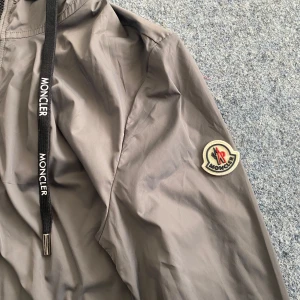 Grå vindjacka från Moncler - Säljer en stilren grå vindjacka från Moncler med dragkedja och justerbar huva. Jackan har Moncler-logotypen på ärmen och svarta dragsnören med märkesdetaljer. Perfekt för blåsiga dagar!