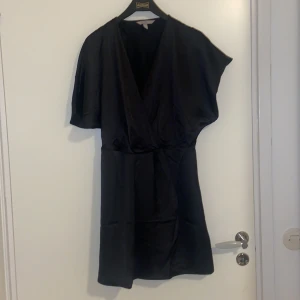 Svart omlottklänning från H&M - Elegant svart omlottklänning från H&M med korta ärmar och v-ringning. Perfekt för en stilren look. Klänningen har en mjuk och följsam passform som ger en smickrande siluett.