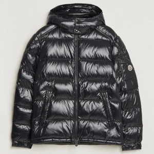 Svart pufferjacka från Moncler - Snygg svart pufferjacka från Moncler med glansig finish och huva. Jackan har en dragkedja framtill och Moncler-logga på ärmen. Insidan har ett unikt serietidningsmönster. Jag säljer den för den va för stor för mig. Den är i st S men känns som M. Aldrig använd 