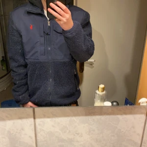  Ralph Lauren jacka - Säljer en snygg mörkblå fleecejacka från Ralph Lauren med dragkedja och broderad logga på bröstet. Jackan har en kombination av fleece och syntetmaterial, vilket gör den både varm och stilren. Perfekt för kyliga dagar! Sitter helt ok på mig som är 171 lite sliten på fleecet vid armbågarna inget som syns man kan bara känna det lite