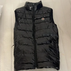 Svart dunväst från Moncler - Snygg svart dunväst från Moncler med glansig yta och dragkedja framtill. Västen är tuekgjord så priset är som det är😊oavsätt är Västen vädligt fin och sitter bra på folk som har L eller M. QR i jackan FUNKAR! som sakt är en väldigt snygg och bra väst för att fånga ögon🥱 Kontakta mig om du har några funderingar//mvh