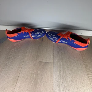 Adidas Predator fotbollsskor - Säljer ett par Adidas Predator fotbollsskor i blått och orange med vita detaljer. Skorna har snörning och är designade för optimal prestanda på planen. Perfekta för fotbollsspelare som vill ha stil och funktion.