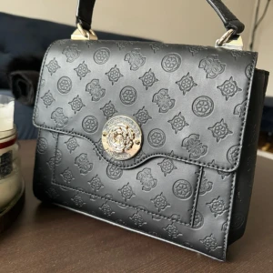 Svart handväska från Guess - Snygg svart handväska från Guess med präglat mönster och en elegant guldlogga på framsidan. Väskan har en praktisk storlek med ett rymligt inre och flera fack. Perfekt för att bära dina nödvändigheter med stil.