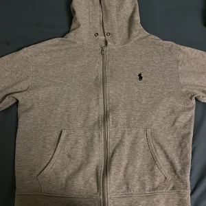Grå hoodie från Ralph Lauren - Säljer en grå hoodie från Ralph Lauren med dragkedja och klassisk logga på bröstet. Tröjan har en bekväm passform och praktiska fickor framtill.