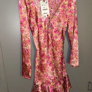Blommig klänning med volang, slutsåld oanvänd Zara  - Säljer en färgglad blommig klänning med volangdetaljer och v-ringning. Klänningen har långa ärmar och en härlig mix av rosa och orange nyanser. Perfekt för att lysa upp garderoben!
