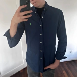 Mörkblå polo skjorta - Säljer en stilren mörkblå skjorta från Ralph Lauren i mycket bra skick. Skjortan har långa ärmar och knappar framtill samt en liten broderad logga på bröstet. Perfekt för både vardag och fest!                  Priset är inte hugget i sten🙌