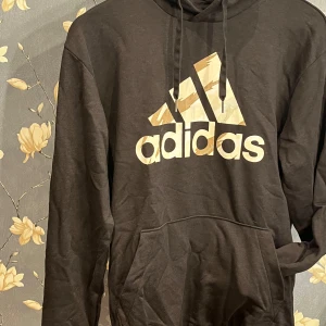 Svart hoodie från Adidas - Säljer en snygg svart hoodie från Adidas i storlek S. Den har en stor logga på framsidan i guld och en praktisk ficka framtill. Perfekt för höst och vinter, och den är superbekväm att ha på sig. Passar både till träning och vardags. 🖤