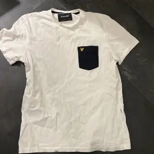 Säljer en stilren vit t-shirt från Lyle & Scott i bomull. Den har en snygg svart bröstficka med deras ikoniska gula logga. Perfekt för vardagsbruk eller en avslappnad dag ute. Kortärmad och i bra skick!