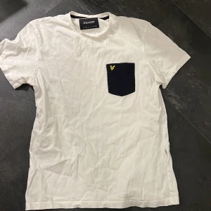 Vit t-shirt med ficka från Lyle & Scott - Säljer en stilren vit t-shirt från Lyle & Scott i bomull. Den har en snygg svart bröstficka med deras ikoniska gula logga. Perfekt för vardagsbruk eller en avslappnad dag ute. Kortärmad och i bra skick!