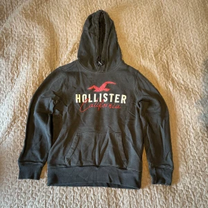 Svart hoodie från Hollister - Säljer en svart hoodie från Hollister i storlek XS. Den har en klassisk design med röd och vit logga på bröstet och en praktisk magficka. Perfekt för en avslappnad stil och passar bra till både höst och vår.
