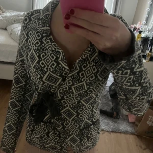 Svartvit mönstrad kofta med knytband - Säljer en snygg svartvit kofta med geometriskt mönster. Den har långa ärmar och ett knytband i midjan för en fin passform. Perfekt för höst och vinter, både mysig och stilren. Passar bra till både vardag och fest!