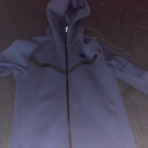 Nike Tech mörkblå - Säljer en blå kofta från Nike i bra skick. Den har en dragkedja framtill och en justerbar huva. Perfekt för höst och vår, med en stilren design och Nike-logga på bröstet. Passar både till träning och vardag.