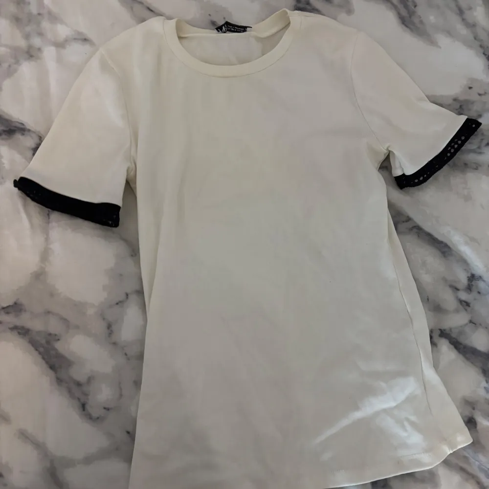 Säljer en stilren vit t-shirt från Zara i storlek XS. Den har korta ärmar med svarta detaljer vid ärmsluten som ger en snygg kontrast. Perfekt för en avslappnad vardagslook eller att styla upp med accessoarer.. T-shirts.