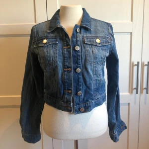 Blå jeansjacka - Säljer en snygg blå jeansjacka i mycket bra skick. Den har en klassisk design med knappar framtill och två bröstfickor med knappdetaljer. Jackan är croppad och har långa ärmar, perfekt för vår och sommar. Passar både till vardags och fest! Använd få gånger!