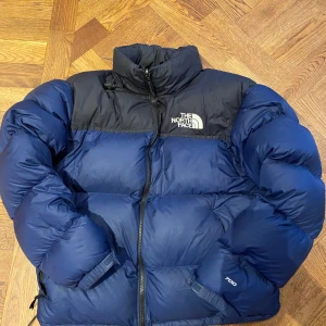 Blå pufferjacka från The North Face - Säljer en blå pufferjacka från The North Face i mycket bra skick. Jackan har en dragkedja framtill och är perfekt för kalla vinterdagar. Den är långärmad och har en snygg logga på bröstet. Supervarm och skön att ha på sig! 