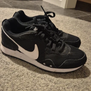Svarta Nike sneakers - Säljer ett par svarta Nike sneakers i bra skick. De har en vit Nike-logga på sidan och snörning framtill. Perfekta för vardagsbruk eller träning. Skorna har en bekväm passform och är gjorda i syntetmaterial.