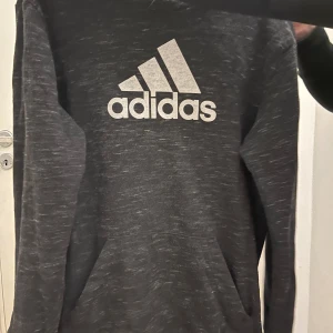 Mörkgrå hoodie från Adidas - Säljer en snygg mörkgrå hoodie från Adidas med deras klassiska logga i vitt på bröstet. Perfekt för både träning och vardag. Den har en skön passform och är i mycket bra skick. Hör av dig vid intresse!