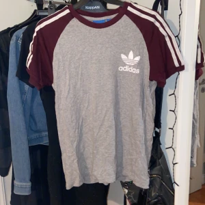 Grå och vinröd t-shirt från Adidas - Säljer en snygg grå t-shirt från Adidas med vinröda ärmar och vita ränder på axlarna. T-shirten är i bra skick💕