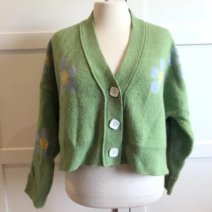 Grön kofta med blommor - Superfin grön kofta med glada blommor i vitt och gult. Den går ner till höften och har stora vita knappar framtill. Perfekt året om, och ger en härlig retrovibe. Ärmarna är långa och koftan är i mycket bra skick. Mjuk och skön.