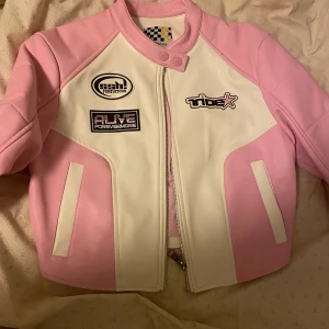 Rosa och vit jacka från Bershka - Säljer en supercool rosa och vit jacka från Bershka. Den har snygga patches med texten 'ride' och 'alive forevermore'. Jackan är i syntetmaterial och har en dragkedja framtill. Perfekt för en trendig look!