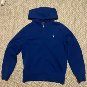 Blå hoodie från Ralph Lauren - Säljer en snygg blå hoodie från Ralph Lauren. Den har en dragkedja framtill och en liten logga på bröstet. Perfekt för kyliga dagar och passar till det mesta. Skön och stilren design!