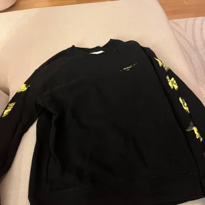 Svart sweatshirt från Off-White - Säljer en svart sweatshirt från Off-White med coola gula detaljer. Tröjan har ett stort grafiskt tryck på ryggen och matchande mönster på ärmarna. Perfekt för en streetwear-look! 🖤💛